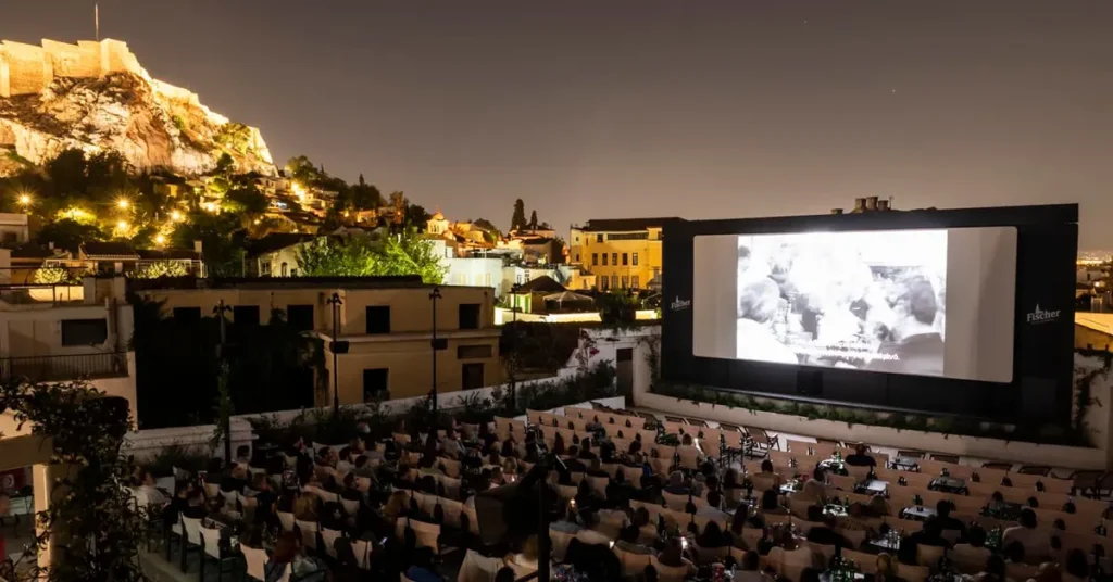 People watching a movie at the open-air cinema Cine Paris with the illuminated Acropolis visible above them.