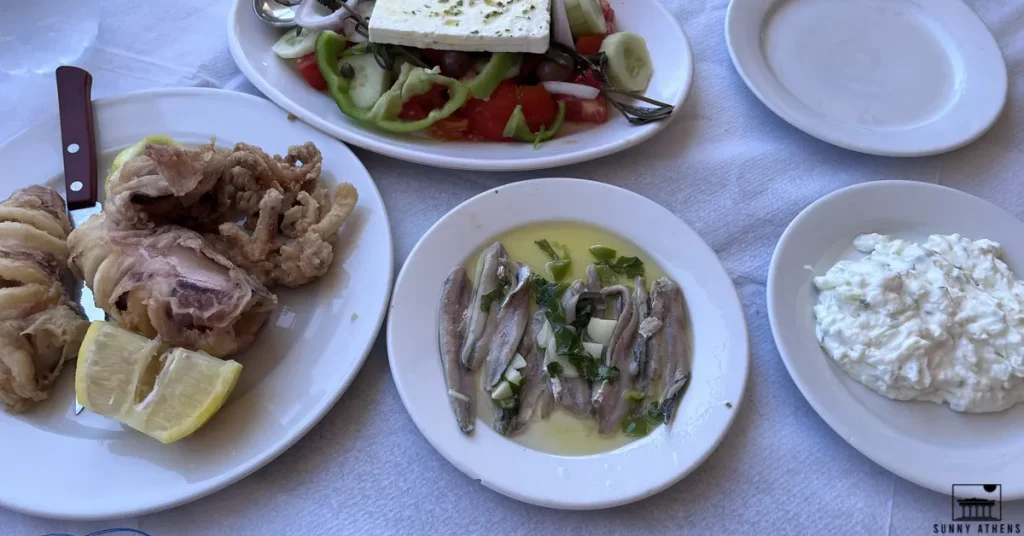 A table set with a variety of Greek dishes including fried calamari, a Greek salad with feta, marinated anchovies, and tzatziki.