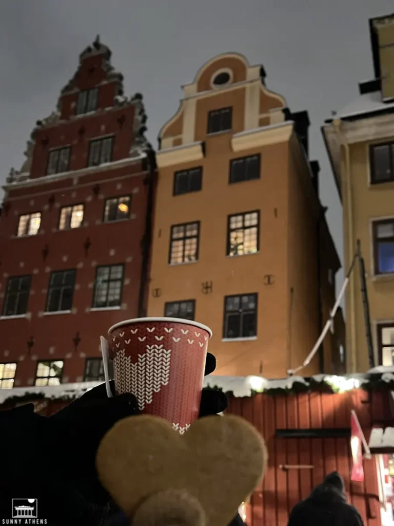 Stockholm Neighborhood Guide for First-Timers: My hand holding a red cup of coffee and a heart-shaped cookie, with historic colorful buildings in the background at Stortorget.