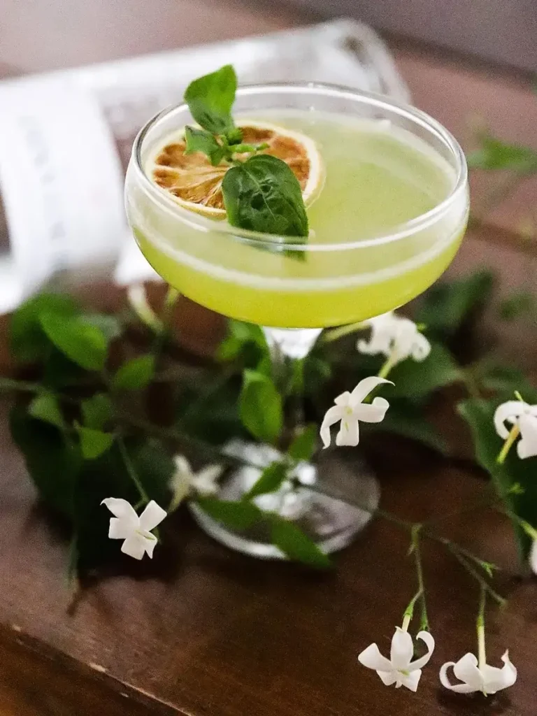 A glass with a green cocktail in it, decorated with a slice of lime and two basil leaves. 