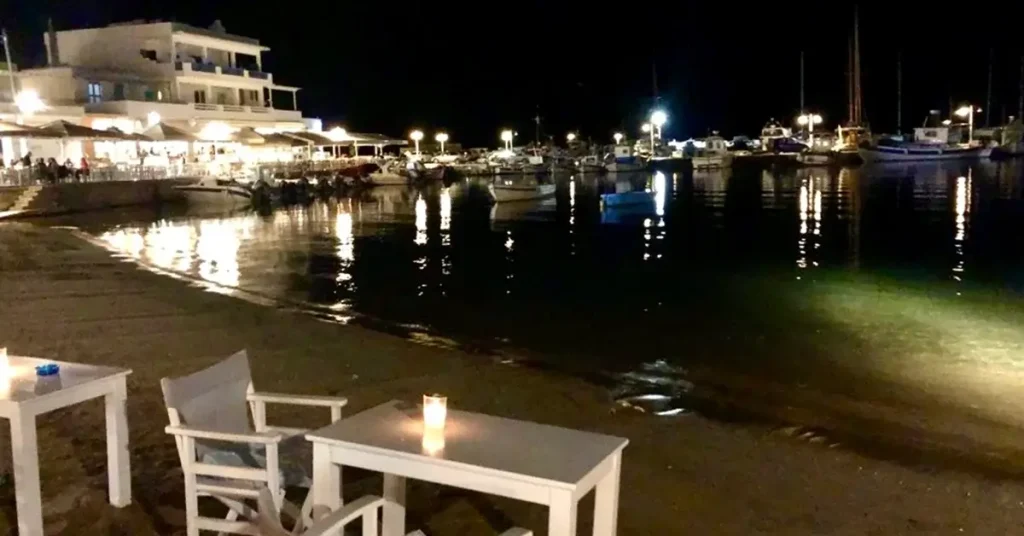 A cozy bar by the sea at night in Piso Livadi, part of Paros Greece nightlife, with small boats and tavern lights twinkling in the background.