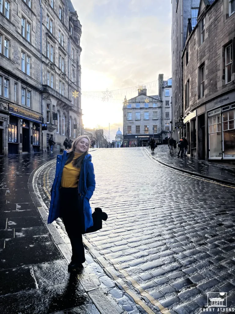 4 days in Edinburgh itinerary: Chrysavgi smiling on a wet cobblestone street with sunlight behind.