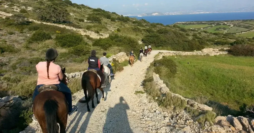 People riding horses in a downhill dirt road in Paros Greece with the Aegean Sea in the background.