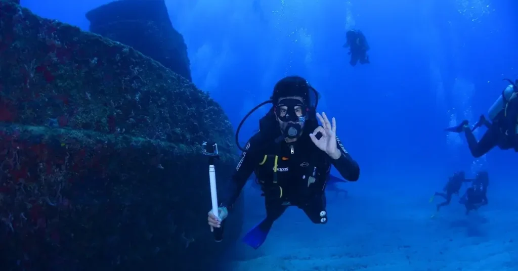 Hidden gems in Paros, Greece: a diver giving an OK signal with one hand and holding a measuring device in the other, next to a shipwreck.