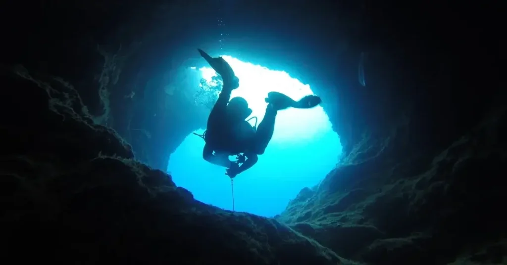 A scuba diver swimming in a cave underwater with sunlight showing from an opening above.