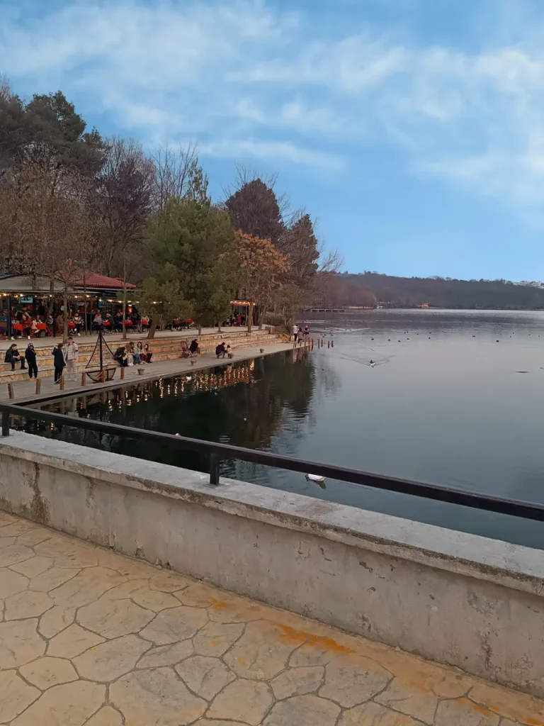 The Grand Park of Tirana lake, featuring ducks in the water and people sitting on a platform at the shore.