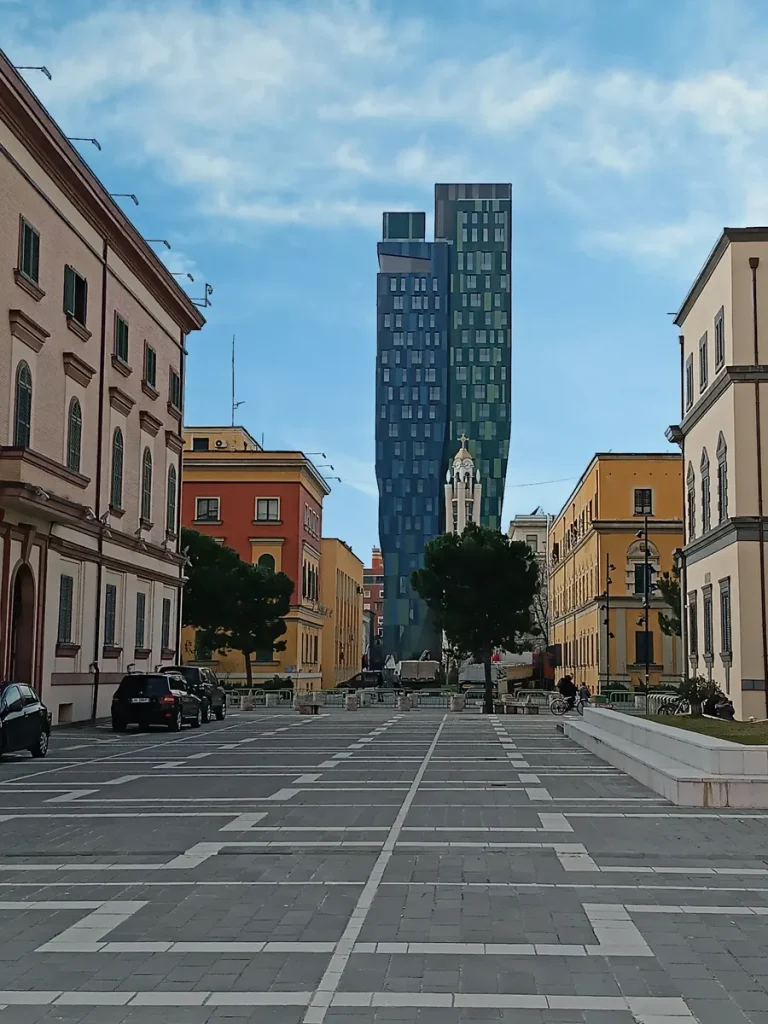 A paved city street in Tirana with a tall, oddly-shaped blue skyscraper in the background.