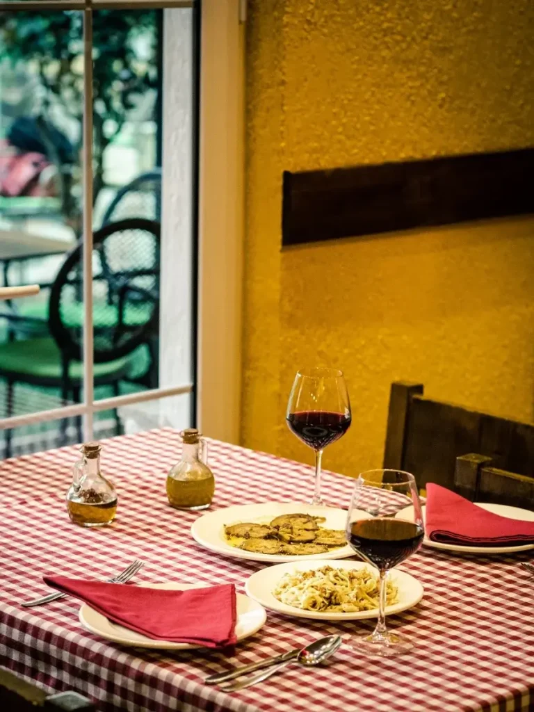 Two pasta dishes and two glasses of red wine on a table with a white and red square tablecloth in a trattoria.