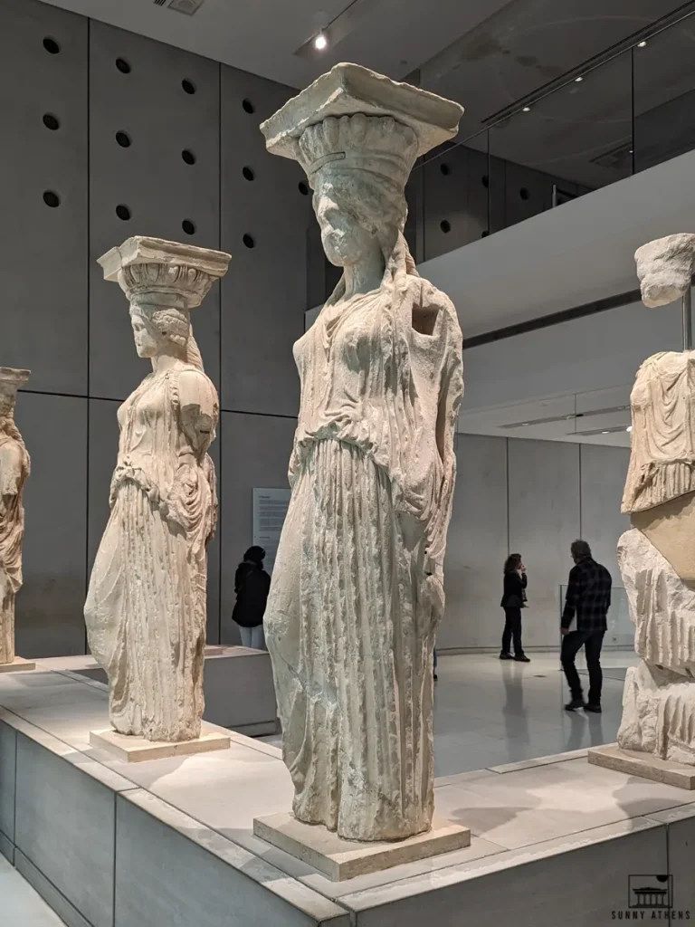 Ancient Caryatid statues on display at the Acropolis Museum, with visitors walking in the background.