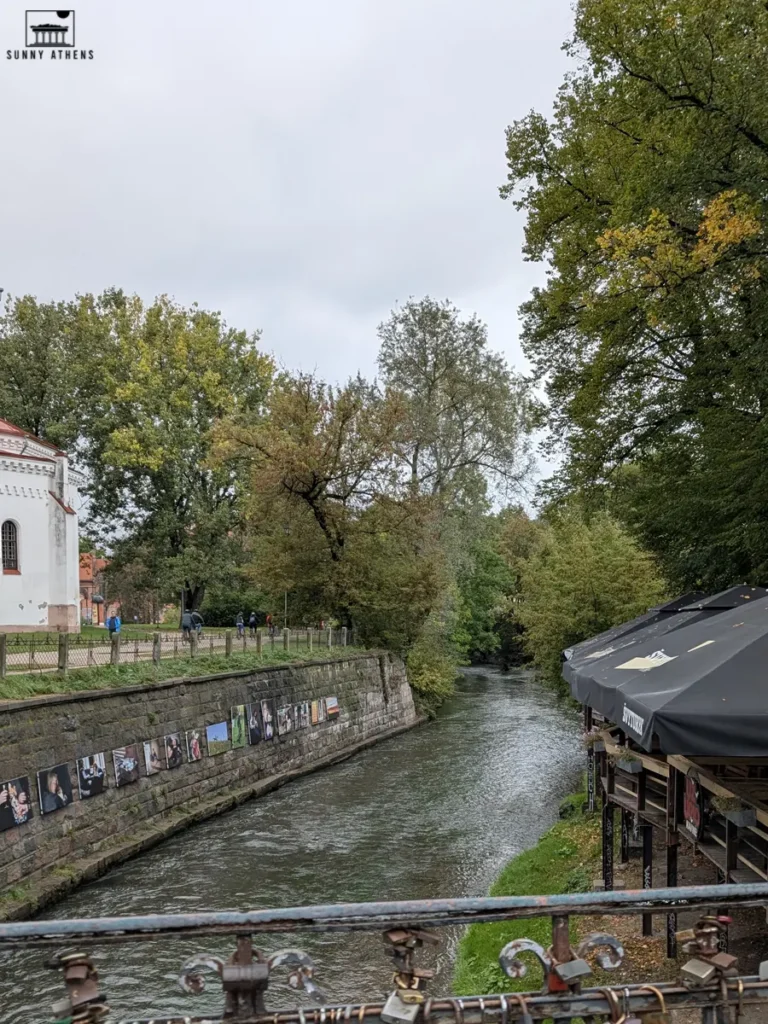 3 days itinerary of Vilnius: the calm river and green trees of Uzupis, with photo art displayed along the wall by the water.