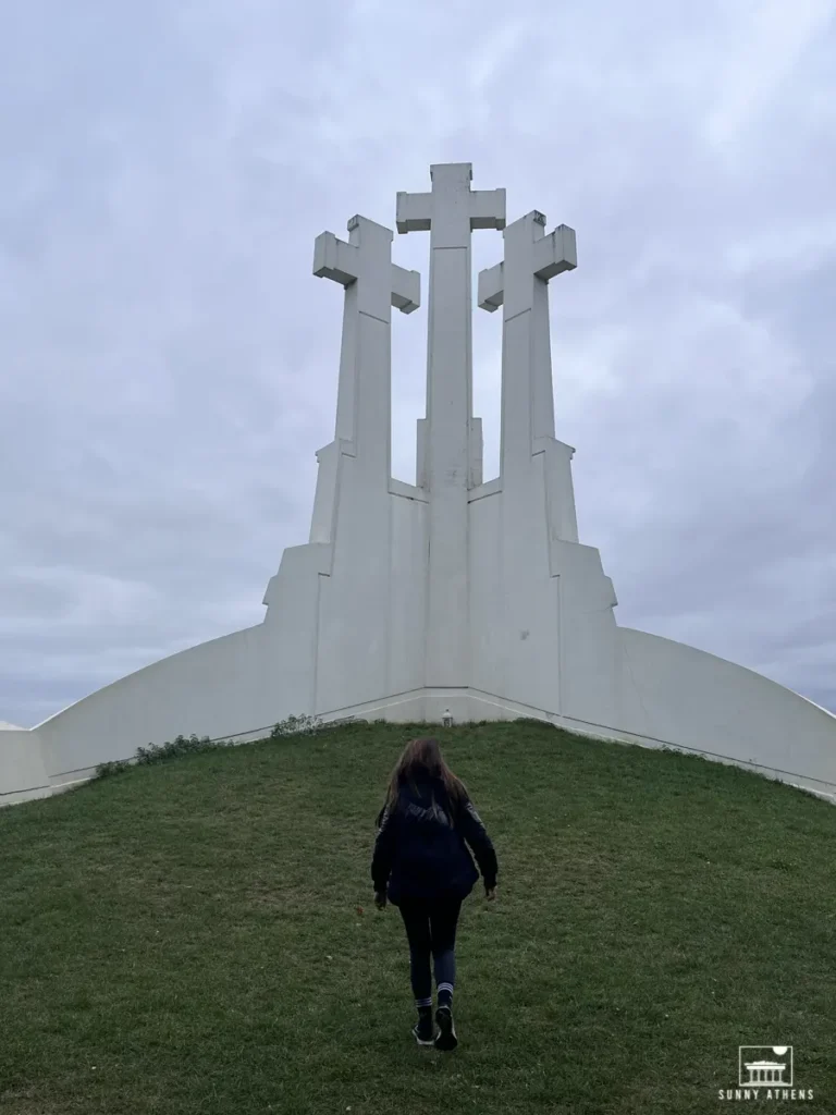 Chrysavgi walking towards the iconic Three Crosses Monument, standing tall against a cloudy sky.