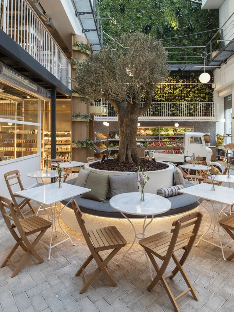 The cozy interior of Ergon House restaurant, featuring an olive tree in the center, marble tables, wooden chairs, and a vertical green wall.