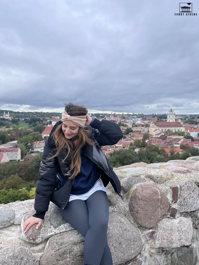 Chrysavgi sitting on a stone wall of Gediminas Castle Tower, with a panoramic view of Vilnius behind her, a highlight of the 3 days itinerary of Vilnius.