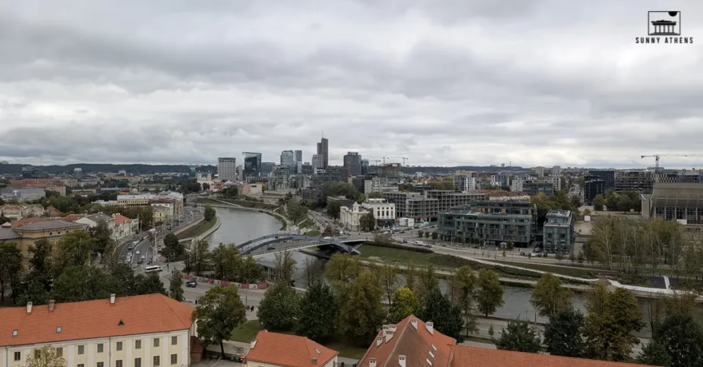 A panoramic riverside view of modern Vilnius with buildings and bridges under a cloudy sky.