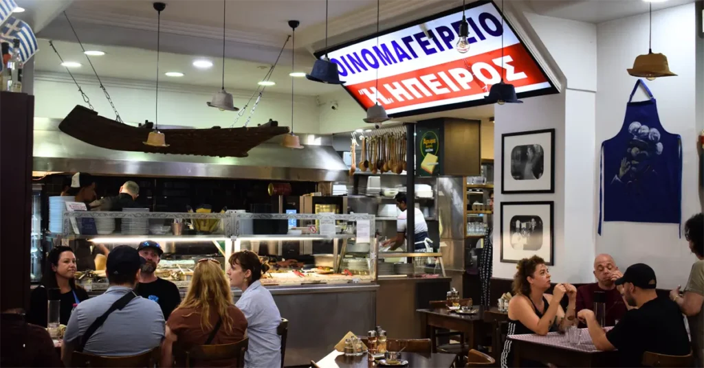 Interior of Oinomageireio Epirus, featuring a bright sign, open kitchen, and people enjoying their meals at wooden tables.