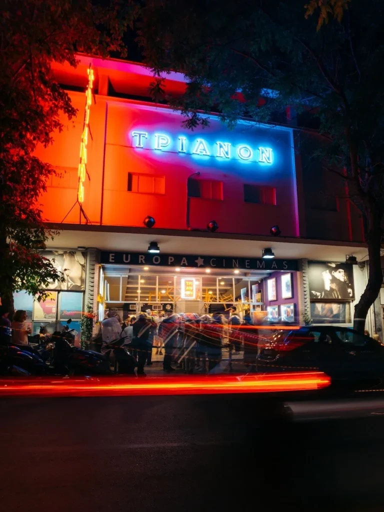 Night streetview of the entrance of the arthouse cinema Trianon in Athens with bright saturated colors coming from the marquee signs and a crowd at the entrance buying tickets