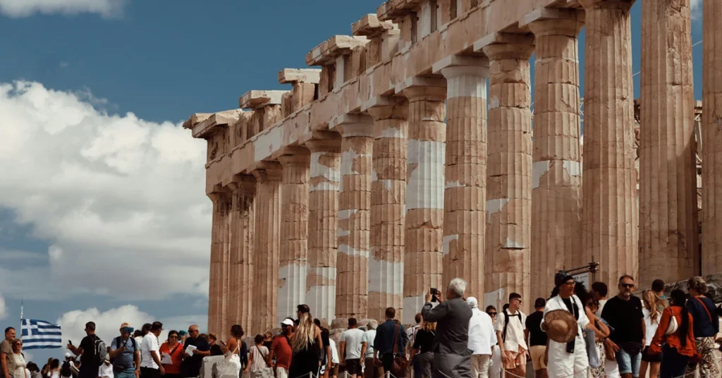 How to escape the crowds in Athens: Tourists walking and photographing the iconic columns of the Parthenon under a sunny sky.