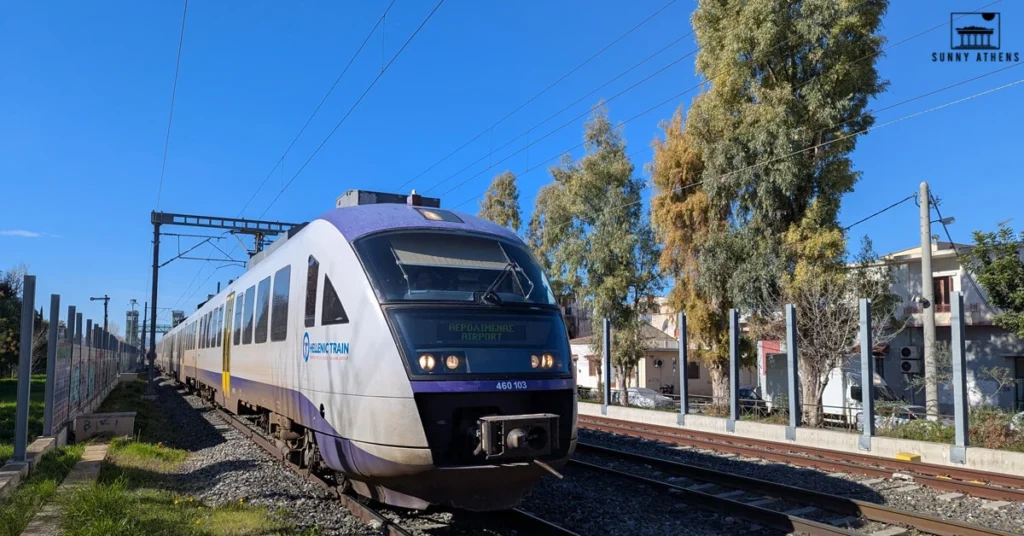 The suburban railway of Athens, labeled "Hellenic Train", approaching a station under a clear blue sky, surrounded by trees and residential buildings.