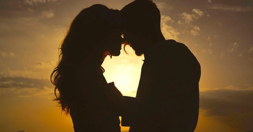 The darkened silhouettes of a man and a woman in front of the sun holding hands and staring each other's eyes from very close