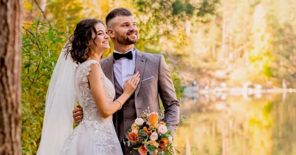 A smiling newlywed couple consisting of a man and a woman holding each other softly in front of a sunlight lake with trees, thinking about their honeymoon in Athens