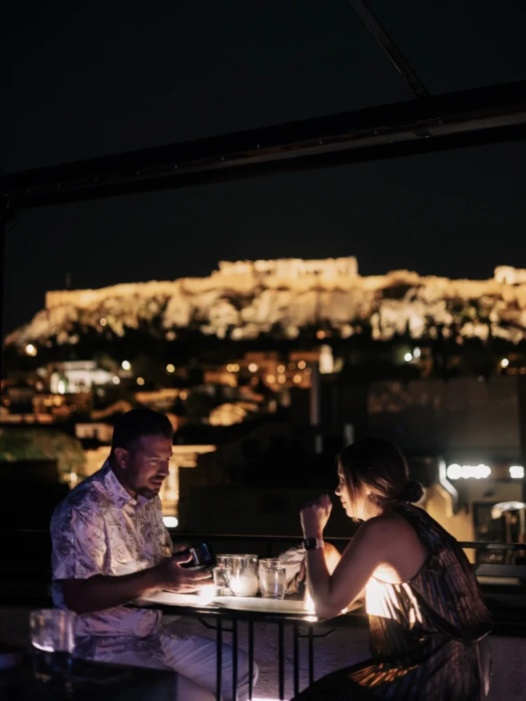 A couple having drinks at the rooftop bar Couleur Locale with a glowing nighttime view of the Acropolis.