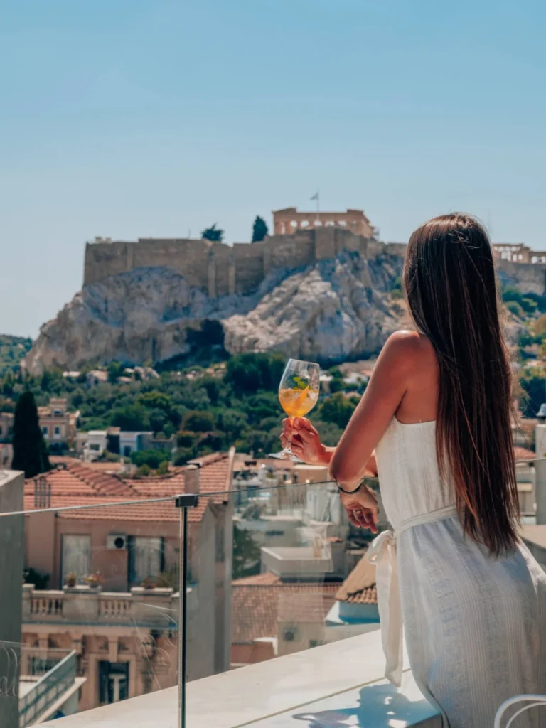 Honeymoon in Athens: A woman in a white dress enjoying a drink on the rooftop of New Hotel Athens with the Acropolis in the background.