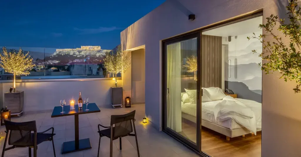 Evening view of a penthouse room and its spacious terrace in Ivis 4 Boutique Hotel in Psiri, Athens, with the illuminated Acropolis in the background.