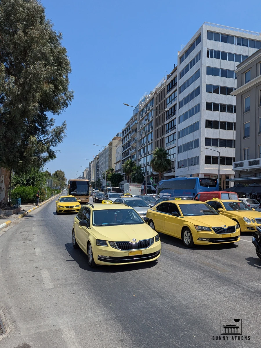 Yellow taxis driving along a busy avenue in central Piraeus on a sunny day.
