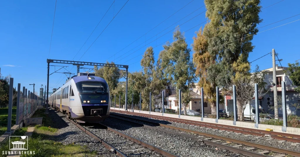 A suburban train arriving at a station near Athens, under clear blue skies.