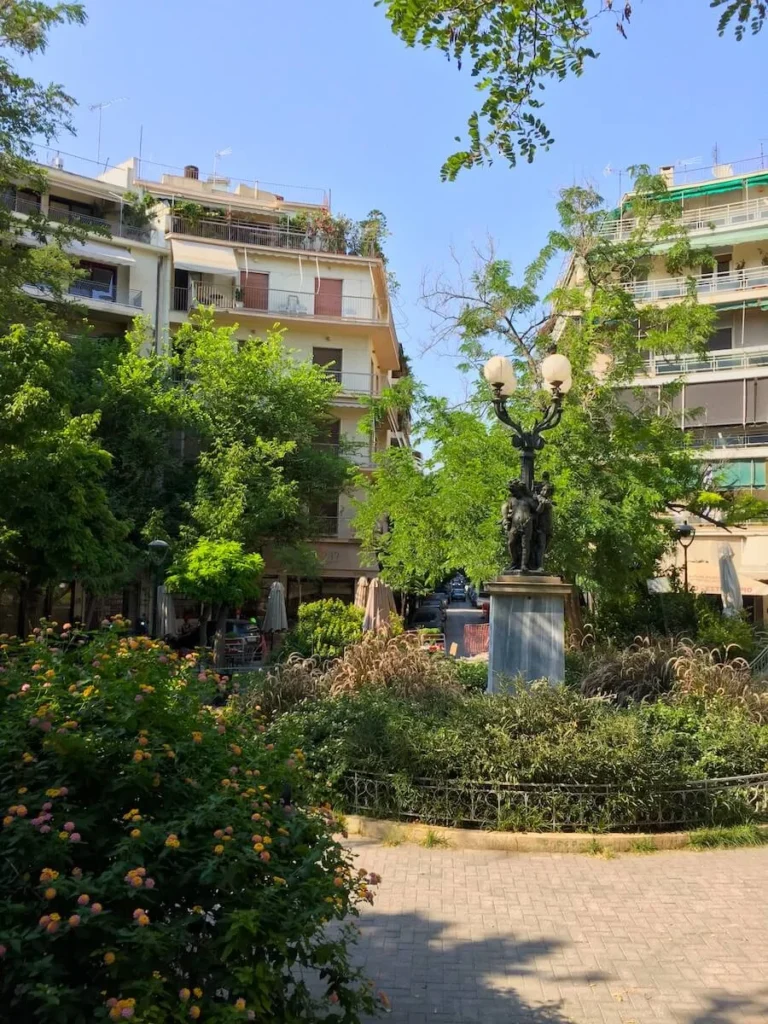 View of Agios Georgios square in Kypseli with trees amd bushes and the statue of the Three Eros in the middle