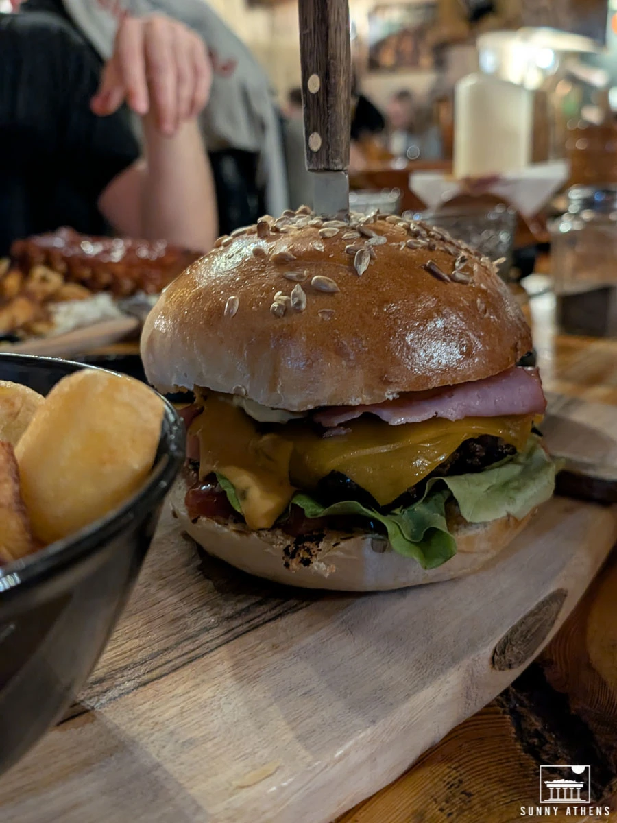 A large burger skewered with a knife and served with a side of potatoes on a wooden board at Excalibur Restaurant.