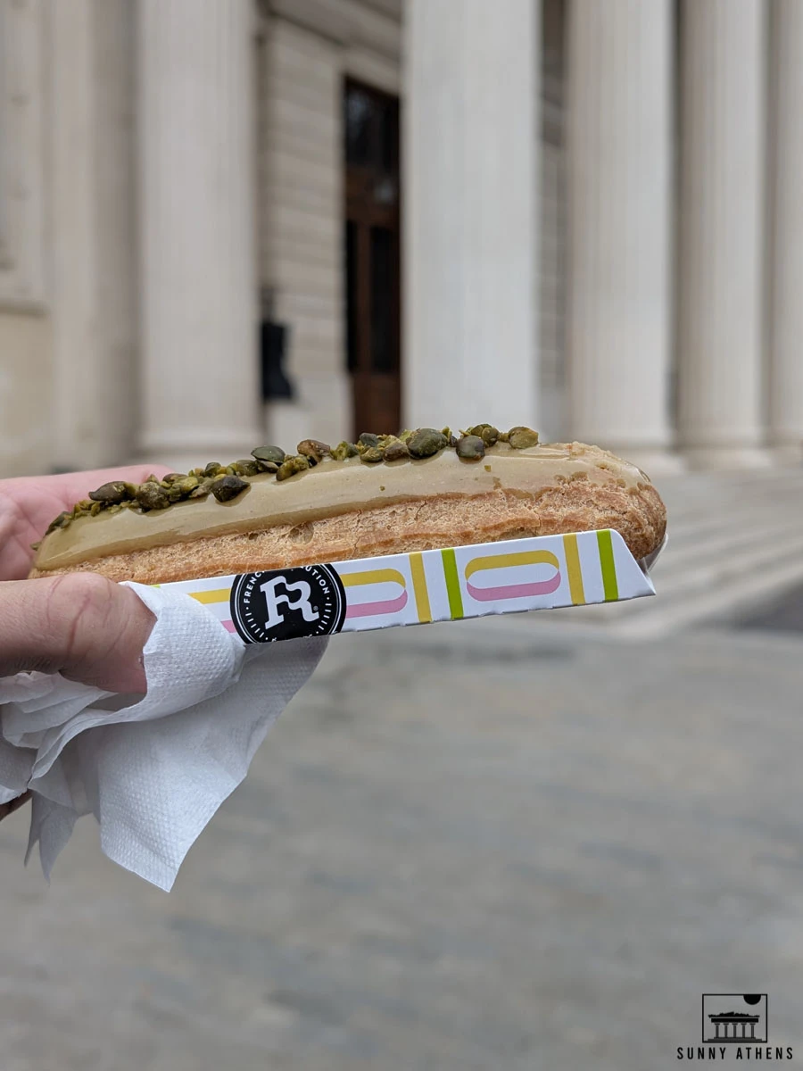 A pistachio eclair from "French Revolution" is held up with the columns of the Romanian Athenaeum in the background.