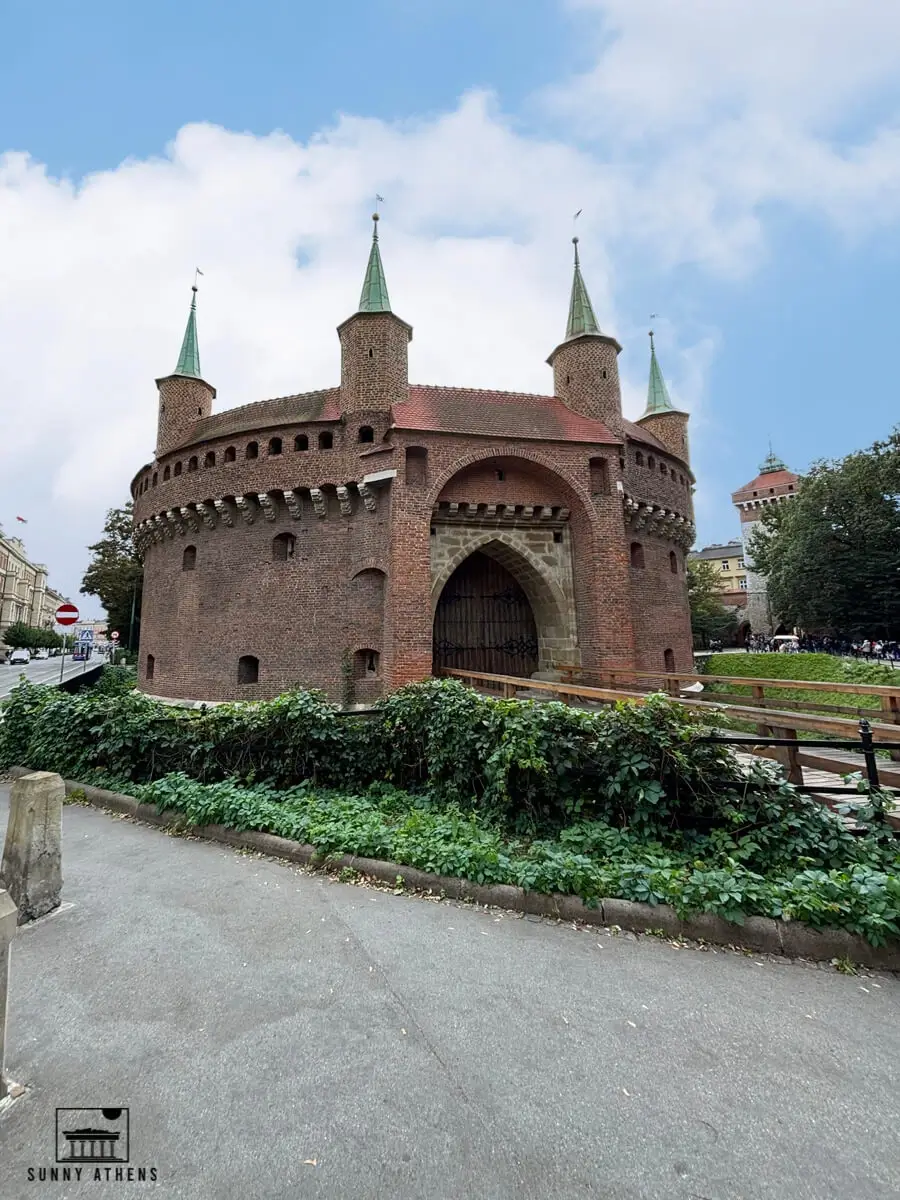 The historic, circular red brick Barbican fortress in Krakow, surrounded by greenery.