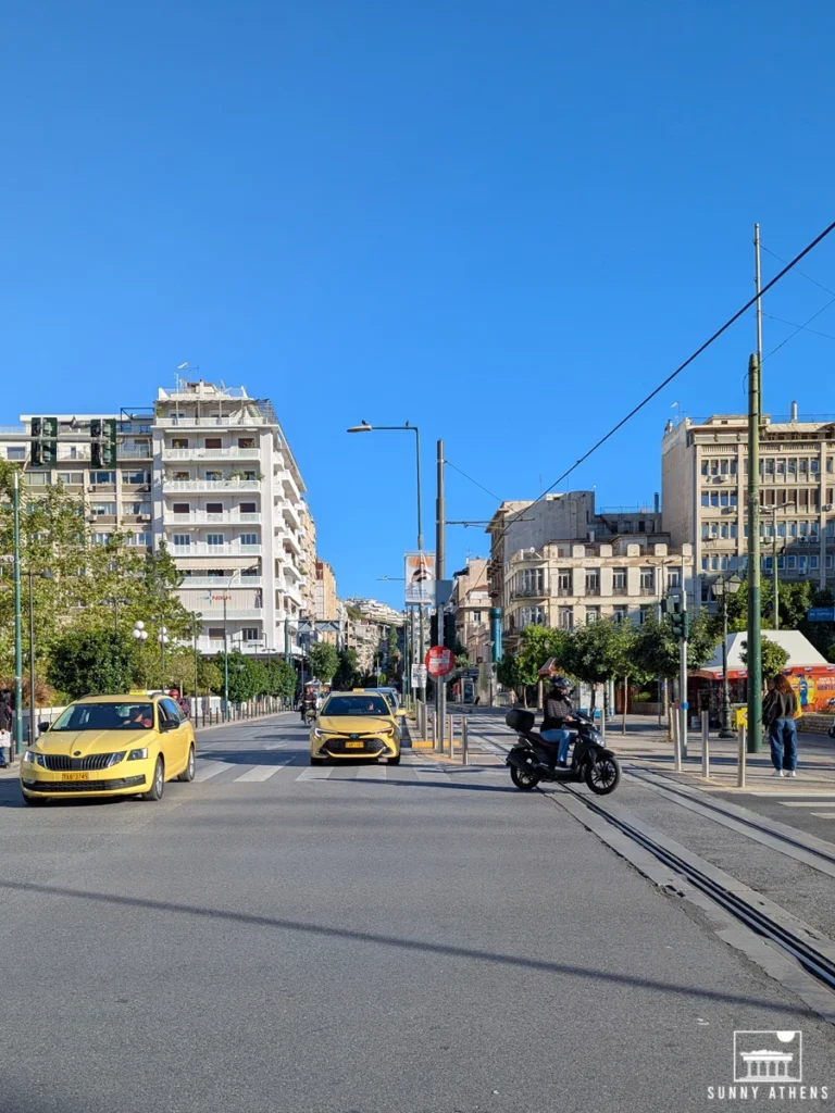 A sunny street scene in Piraeus with apartment buildings, yellow taxis, and a scooter.