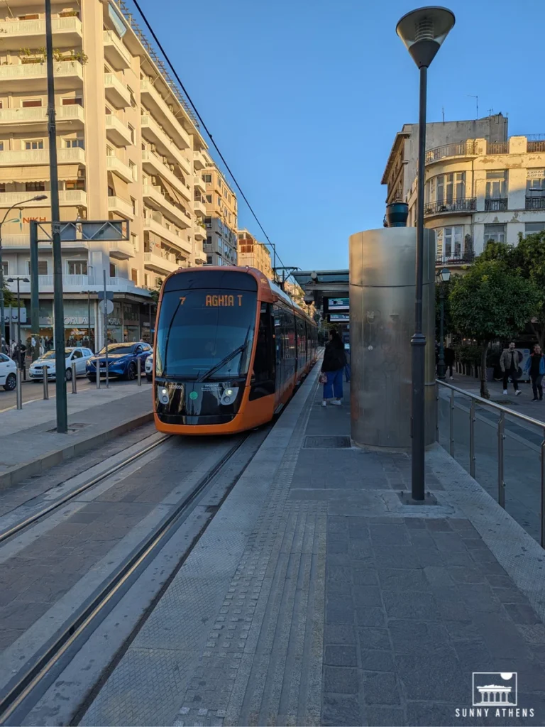 An orange tram arriving at a stop in Piraeus with modern buildings in the background.