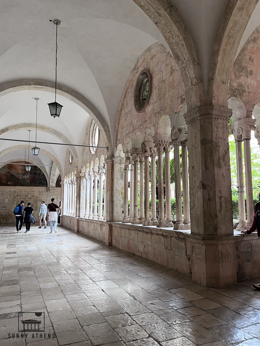 The Romanesque cloister courtyard of the Franciscan Monastery featuring double stone columns, arched walkways, and green trees