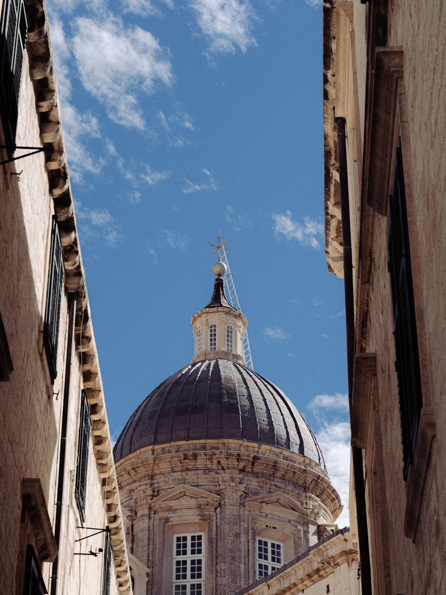The ornate Baroque dome of Dubrovnik Cathedral framed between high stone walls of an Old Town alley under a blue sky