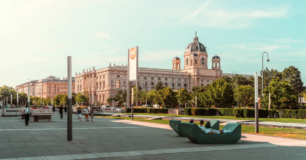 Visitors relaxing on green Enzi lounge seats in the MuseumsQuartier courtyard with the Kunsthistorisches Museum dome in Vienna