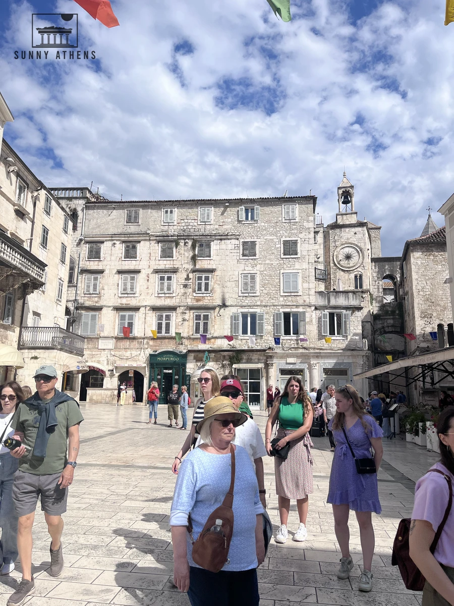 Pedestrians walking across the stone paving of People's Square in front of historic townhouses and the Romanesque clock tower