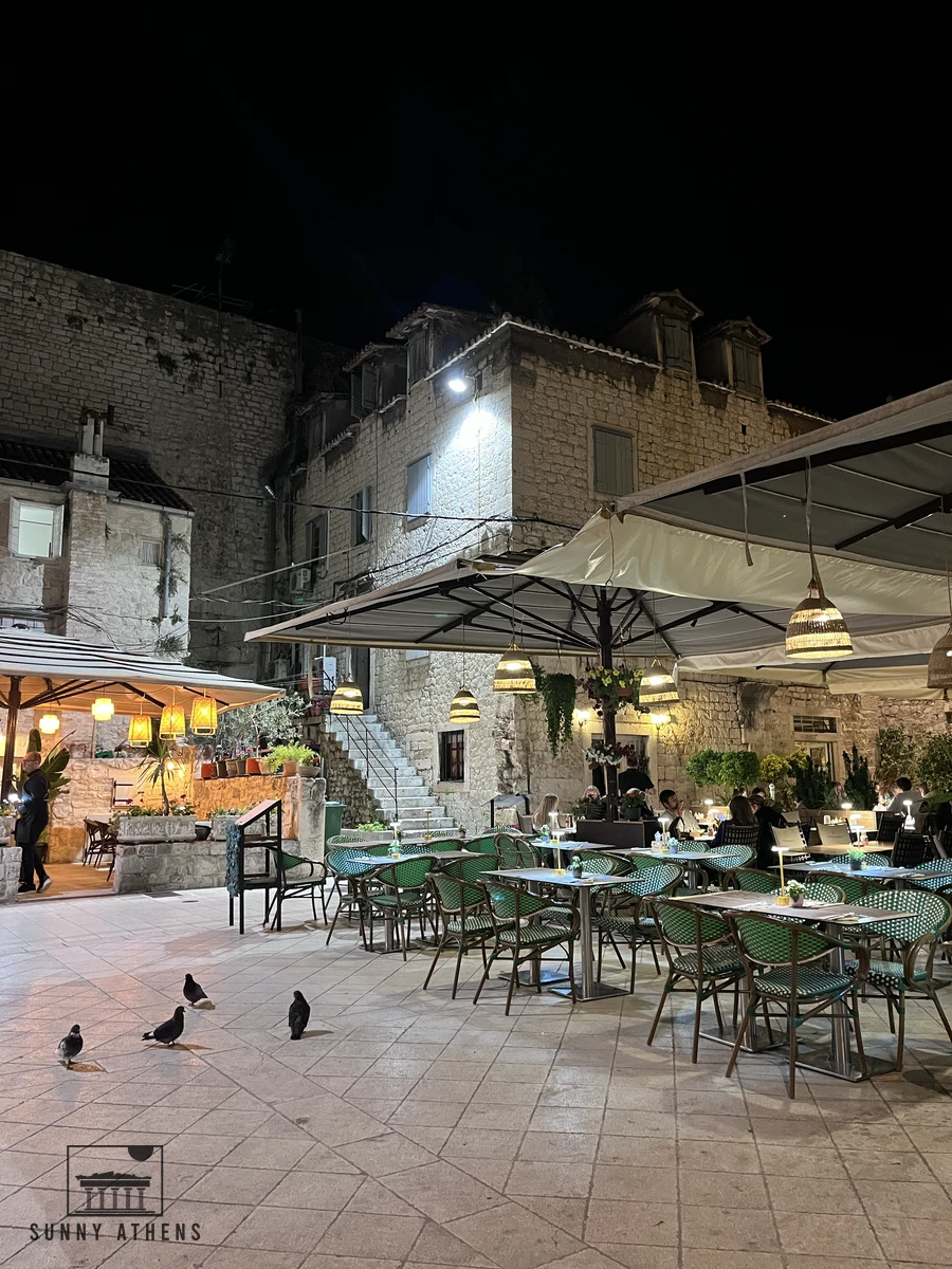 Outdoor restaurant terrace with illuminated tables and green chairs in a historic stone square at night in Split